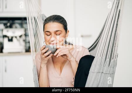 Positive Frau mittleren Alters mit geschlossenen Augen trinken heißes Getränk Während Sie in der Küche in der Hängematte sitzen und moderne Küchengeräte an haben Unscharfer Hintergrund Stockfoto