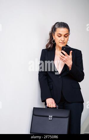 Ernsthafte Unternehmerin mittleren Alters mit Pferdeschwanz in schwarzem Anzug Textnachrichten auf dem Mobiltelefon, während Sie auf weißem Hintergrund mit stehen Ordner Stockfoto