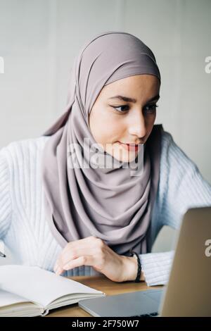 Porträt einer ethnischen Freiberuflerin in Hijab mit Laptop, während Sitzen am Tisch im Café und Arbeiten aus der Ferne Stockfoto