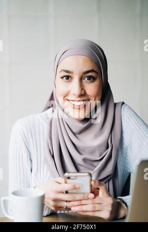 Porträt einer ethnischen Freiberuflerin in Hijab mit Smartphone während Sitzen am Tisch im Café und Arbeiten aus der Ferne Stockfoto