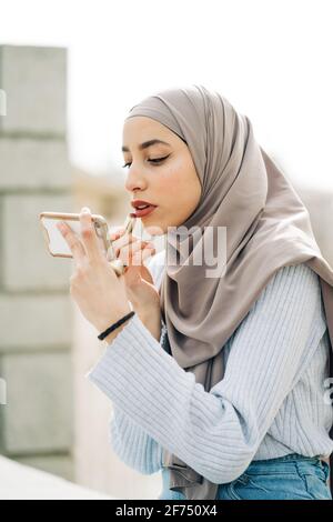 Seitenansicht der muslimischen Frau im Kopftuch mit Lippenstift und Blick auf den Bildschirm des Smartphones, während Make-up auf der Straße Stockfoto