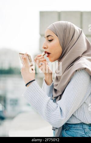 Seitenansicht der muslimischen Frau im Kopftuch mit Lippenstift und Blick auf den Bildschirm des Smartphones, während Make-up auf der Straße Stockfoto
