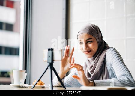 Niedriger Winkel der fröhlichen muslimischen Frau im Kopftuch mit Video Rufen Sie auf dem Stativ ein Smartphone an, um während des Sitzens zu bloggen Tisch im Café Stockfoto