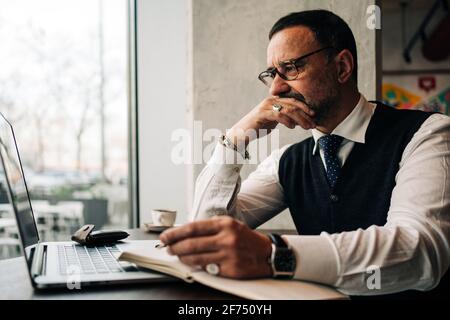 Nachdenklich reif ethnisch männlich exekutive berühren Mund am Tisch mit Netbook und Tagebuch in der Cafeteria Stockfoto