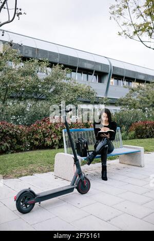 Fokussierte weibliche Unternehmerin sitzt auf der Bank in der Nähe von modernen Elektroroller Und einen Plan im Notebook während eines abgelegen arbeitenden Stadtparks zu schreiben Stockfoto