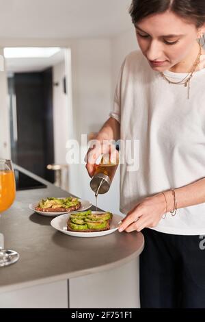 Crop konzentriert junge Frau in legerer Kleidung Zugabe von Olivenöl Auf leckeren Avocado-Toast während der Zubereitung des Frühstücks in der Küche Stockfoto