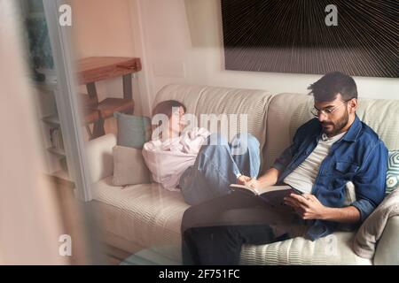 Müde junge Frau in legerer Kleidung, die auf einem bequemen Sofa schläft Beinahe fokussierter ethnischer Freund, der bei Tageslicht Buch liest Stockfoto