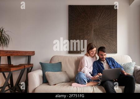 Fröhliches junges, multirassisches Paar in lässigen Outfits, lächelnd im Sitzen Auf dem Sofa und Videogespräche über den Laptop Stockfoto