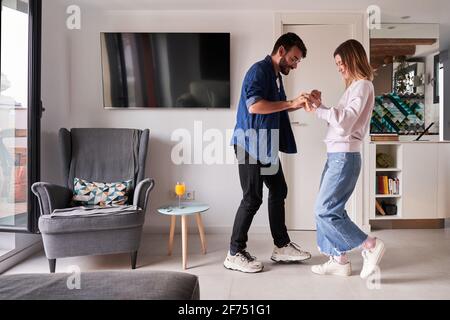 Seitenansicht eines aufgeregt jungen multirassischen Paares in stilvoller Kleidung Hände halten, während sie tagsüber in einer modernen Wohnung zusammen tanzen Stockfoto