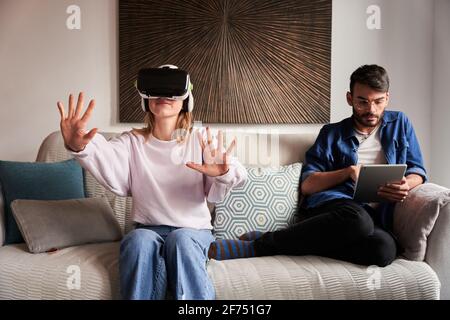 Positive junge Frau erlebt virtuelle Realität in einem modernen Headset, das auf einem bequemen Sofa in der Nähe eines konzentrierten ethnischen Freundes sitzt, der auf einem Tablet surft Stockfoto