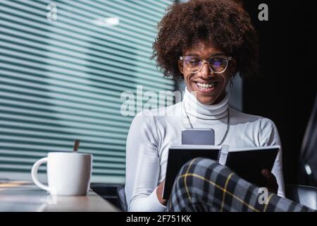 Positive junge ethnische männliche Freiberuflerin in stilvoller Kleidung und Brille Notizen im Planer machen und mit dem Smartphone arbeiten, während Sie aus der Ferne arbeiten Am Tisch w Stockfoto