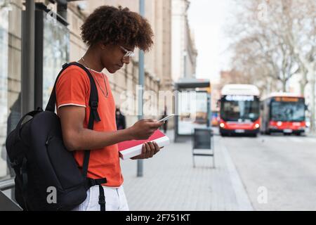 Seitenansicht eines jungen afroamerikanischen Mannes in legerer Kleidung Und Rucksack mit dem Smartphone, während Sie auf der Stadtstraße stehen und Warten auf den Bus Stockfoto