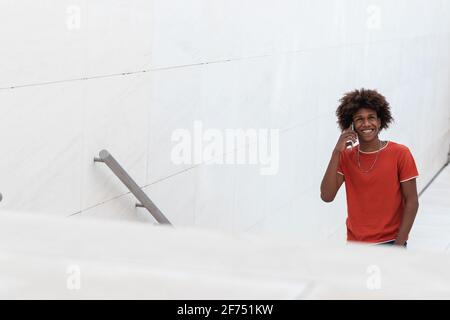 Glücklich junge afroamerikanische Mann Millennial in lässiger Kleidung mit Telefongespräch, während man tagsüber auf der Stadtstraße nach oben ging Stockfoto