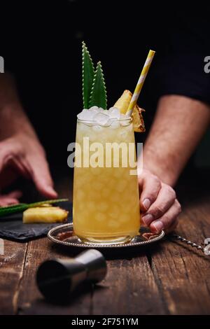Crop anonymen Barkeeper garnieren Alkohol Cocktail mit grünen Blättern und Ananasstück auf Tablett in der Nähe von Schnapsglas am Holztisch Auf schwarzem Hintergrund Stockfoto