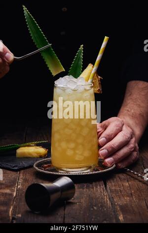 Crop anonymen Barkeeper garnieren Alkohol Cocktail mit grünen Blättern und Ananasstück auf Tablett in der Nähe von Schnapsglas am Holztisch Auf schwarzem Hintergrund Stockfoto