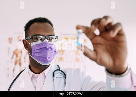 Afroamerikanischer Mann in Uniform mit Stethoskop und verschwommenem Impfstoff In der Hand im Krankenhaus Stockfoto