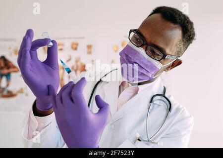 Intelligente afroamerikanische Arzt in medizinische Maske und Handschuhe Füllung Spritze mit blauer Injektionsflüssigkeit im Krankenhaus Stockfoto