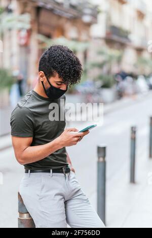 Vertikales Porträt eines jungen lateinamerikaners, der sein Smartphone auf der Straße überprüft. Er trägt lässige Sommerkleidung und eine schwarze Schutzmaske. Stockfoto