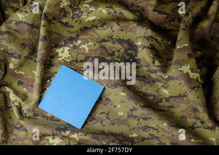 Ein Fragment einer zerknitterten Tarnuniform und einem blauen Blatt Papier. Quadratisches Blatt für Notizen auf einem speziellen Stoff. Armee-Thema. Stockfoto