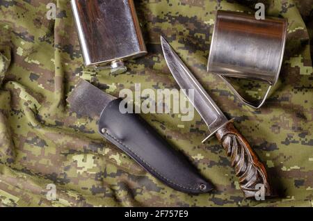 Messer, Scheide, Metallkolben und Becher auf Camouflage-Stoff. Großes Messer mit Holzgriff, schwarze Lederscheide liegt auf Militäruniform. Armee oder hu Stockfoto