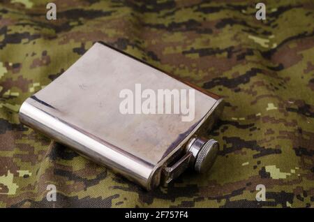 Metallflasche auf Camouflage-Stoff. Glänzend gefärbter Kolben für verschiedene Getränke auf Militäruniformen. Armee-Thema oder Jagd. Selektiver Fokus. Stockfoto
