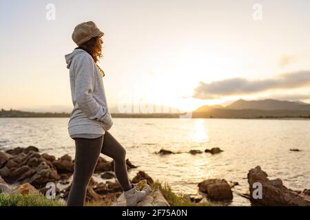 Allein nachdenkliche Frau, die auf Meeresfelsen steht und den Sonnenuntergang oder Sonnenaufgang zwischen den Bergen am Horizont beobachtet. Casual hübsche Person mit Wollhut findet sie Stockfoto