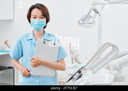 Porträt einer jungen vietnamesischen Krankenschwester in einer medizinischen Maske mit Ordner Beim Stehen im Zahnarztbüro Stockfoto