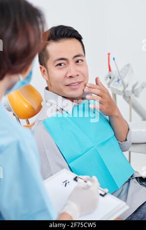 Hübscher junger vietnamesischer Mann, der auf einem Stuhl sitzt und sich darüber beschwert Starke Zahnschmerzen beim Gespräch mit der Krankenschwester Stockfoto