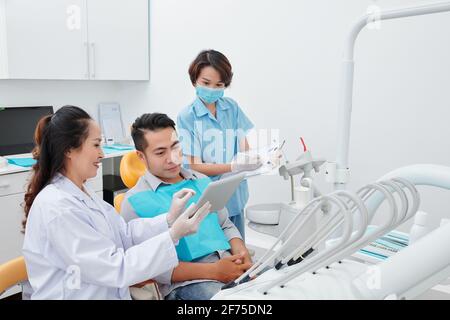 Lächelnder Zahnarzt zeigt dem Patienten einen Tablet-Computer mit Röntgenaufnahme der Zähne Und erklären, was von der Behandlung zu erwarten ist, wenn der Assistent nimmt Hinweise im Dokument Stockfoto