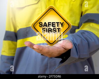 Arbeitsschutzkonzept. Wort „Safety First“-Schild und Hilfssymbol auf quadratischem gelbem Schild auf der Hand von Technikern, die eine gelbe, gut sichtbare Jacke tragen Stockfoto