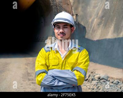 Porträt eines jungen hübschen asiatischen Tunnelbaus in gelber Höhe Visibility Jacke und weißer Schutzhelm stehen mit Armen gefaltet Vorne o Stockfoto