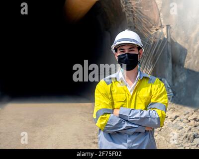 Portrait des jungen asiatischen Tunnelbaus mit einer gut sichtbaren Jacke, Gesichtsmaske und weißem Schutzhelm, der mit gefalteten Armen auf dem eisenbahntunnel steht Stockfoto