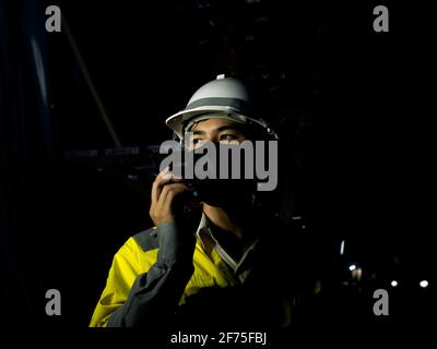 Junger asiatischer Ingenieur mit gelber, gut sichtbarer Sicherheitsjacke, Gesichtsmaske und weißem Schutzhelm oder Schutzhelm mit Walkie Talkie im Dunkeln Stockfoto