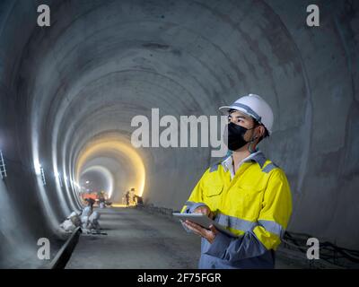 Junge asiatische Tunnel Engineering trägt hohe Sichtbarkeit Jacke, Gesichtsmaske und weißen Schutzhelm arbeiten und mit digitalen Tablet in dunklen Eisenbahn-tunn Stockfoto