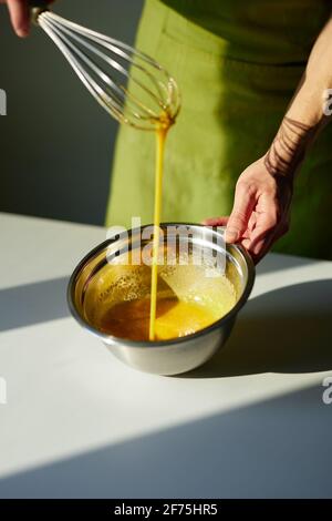 Zubereitung von hausgemachten Kuchen oder Cookie-Konzept. Männchen in grüner Schürze schlägt Zucker und Hühnereier, die mit Drahtbesen und Metallschüssel in eine Tasse geschlagen werden. Vertikales Bild. Hochwertige Fotos Stockfoto