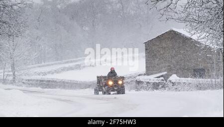 Landwirt auf einem Quad mit Anhänger voller Heu, der eine schneebedeckte Straße entlang fährt, um Schafe zu füttern. Hawes, North Yorkshire, Großbritannien. Stockfoto