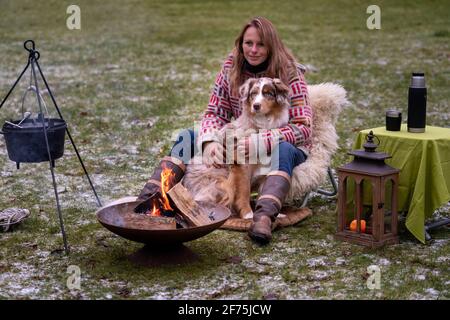 Tricolor Australian Shepherd sitzt neben einer jungen Frau am Lagerfeuer. Im Winter Schnee auf dem Gras. Das Feuer brennt unter dem Kessel Stockfoto