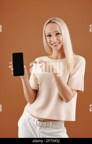 Porträt einer lächelnden jungen hübschen Frau, die auf den Bildschirm zeigt Smartphone in der Hand und Blick auf die Kamera Stockfoto