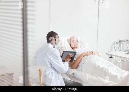 Rückansicht Porträt eines afroamerikanischen Arztes mit digitalem Tablet, während er von einem älteren Patienten im Krankenhausbett sitzt, Platz kopieren Stockfoto