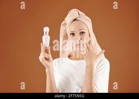 Porträt einer aufgeregten jungen Frau mit einem Handtuch auf dem Kopf Zeigt eine Flasche neuen Parfums oder feuchtigkeitsspendenden Serums Stockfoto