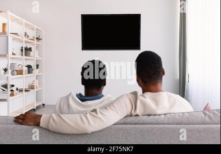 Schwarzes Paar, das mit Mockup Fernsehen schaut und auf der Couch sitzt Stockfoto