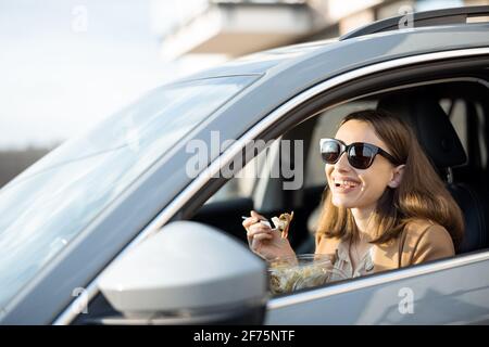 Schöne Frau, die im Auto einen heidnischen Salat isst. Ich habe eine Bestellung zum Essen erhalten. In der Mittagspause haben Sie einen schnellen Snack. Stockfoto