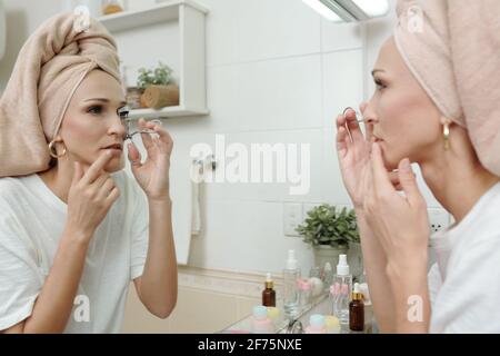 Frau, die auf Badezimmerspiegel schaut, wenn sie Wimpern-Lockenstab vor verwendet Mascara auftragen Stockfoto