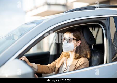 Junge Frau, die mit einer Schutzmaske und einer Sonnenbrille im Gesicht Auto fährt. Gesundheitswesen, Virenschutz, Allergieschutzkonzept. Stockfoto