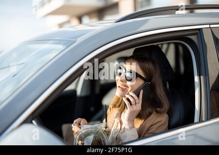 Schöne Frau, die im Auto einen heidnischen Salat isst und am Telefon spricht. Ich habe eine Bestellung zum Essen erhalten. In der Mittagspause haben Sie einen schnellen Snack. Stockfoto