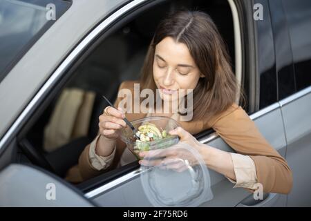 Schöne Frau, die im Auto einen heidnischen Salat isst. Ich habe eine Bestellung zum Essen erhalten. In der Mittagspause haben Sie einen schnellen Snack. Stockfoto