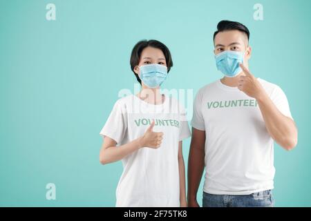 Junges asiatisches Paar zeigt auf Schutzmasken und zeigt Daumen nach oben Bei der Freiwilligenarbeit zusammen Stockfoto