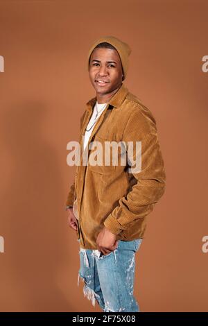 Junger Mann mit Hut lächelnd im Studio vor braunem Hintergrund. Stockfoto