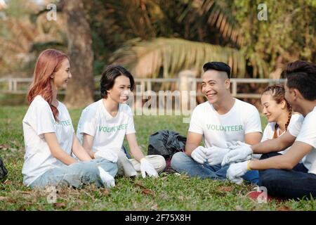 Gruppe lächelnder junger Männer und Frauen, die auf Gras sitzen Im Stadtpark und diskutieren Freiwilligenarbeit Stockfoto