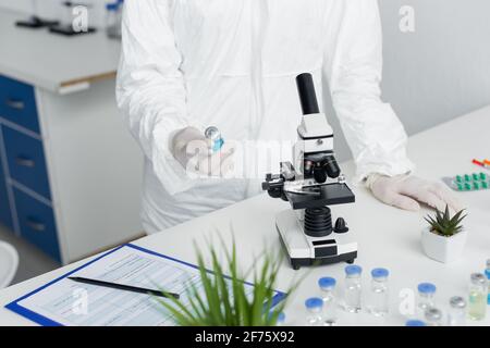 Beschnittene Ansicht eines Wissenschaftlers in einem Hazmat-Anzug, der den Impfstoff in der Nähe hält Mikroskop und Zwischenablage Stockfoto